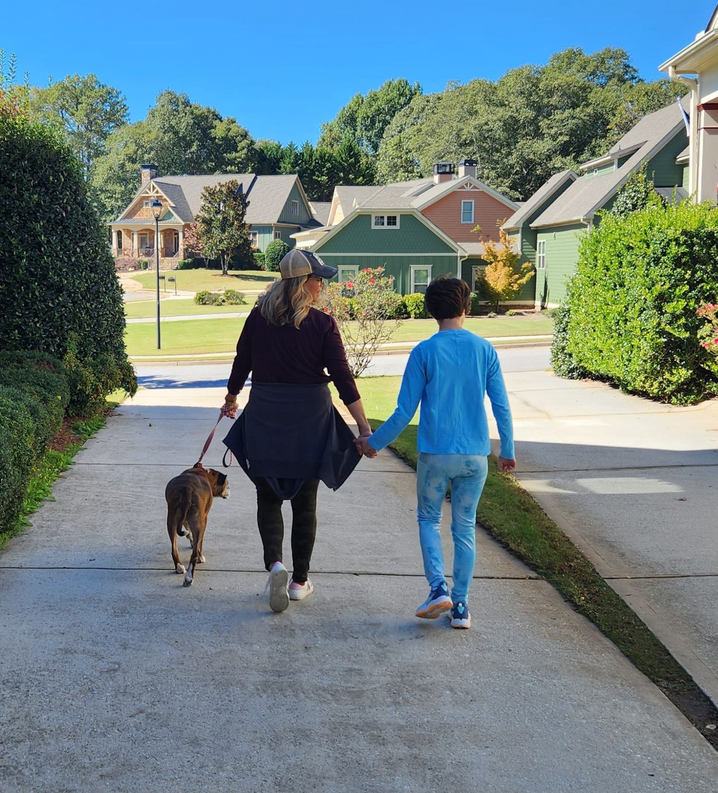 A mother and her daughter walk down a sunny neighborhood street, holding hands, their dog beside them.